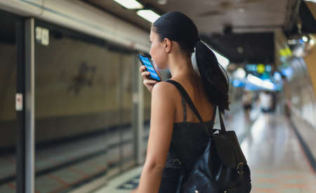 Side view of beauiful woman using mobile phone and talking on the speaker while stand waiting for train to arrive in metro subway. People lifestyle and daily life,commuter and public transport conceptの写真素材