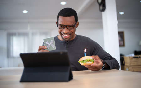 Happy Black man sitting at the table celebrating birthday through video call virtual party with friends at home. Lits and blows out candle on burger and holding gift box. Concept of distance relationsの写真素材