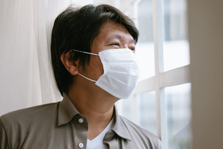 Lonely Korean man in medical facial  mask looking through the window. solation at home for self quarantine. Unhappy male in facemask suffer from coronavirus. Quarantine, corona pandemic concept.の写真素材