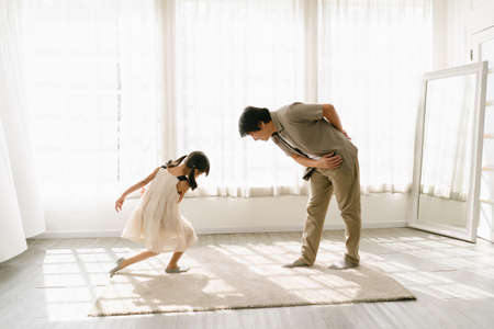 Smiling Asian father standing next to window with invite cute little daughter for dancing waltz at home. Happy loving chinese or japanese dad enjoy time with little girl child in princess skirt.の写真素材