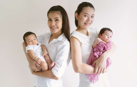 Portrait of two Asian Mothers holding newborn babies are standing back to back and smiling looking at camera isolated on light beige background. Family, Friend, People and child health care concept.の写真素材