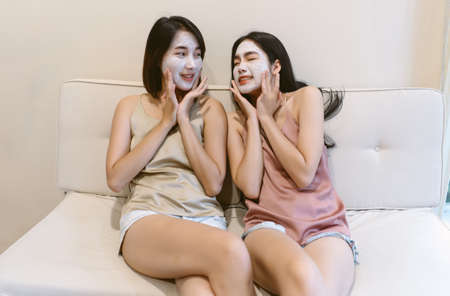 Happy two Asian girls friends relaxing with facial masks while sitting on sofa in living room at home. Skin care and treatment, facial cosmetic mask, natural beauty and cosmetology concept.の写真素材