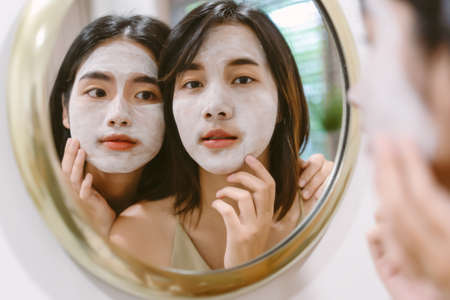 Asian Two beautiful and cheerful girls during applying facial mask treatment looking in mirror in bathroom. Lesbian couple relax and enjoy beauty facial treatment together at home. Skin care conceptの写真素材