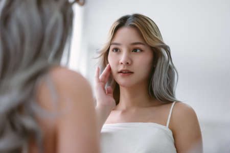 Beautiful young asian woman looking at mirror touching her skin enjoying treatment for skin. Korean or Japanese influencer girl with moisturized soft skin condition, enjoying healthy skin care.の写真素材