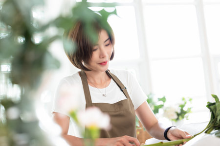 Smiling young Asian Woman Florist small business flower Shop Owner. Influencer beautiful Asian girl vlogger making bouquet flower at table in her workshop at store. Floristry and Flower shop conceptの写真素材