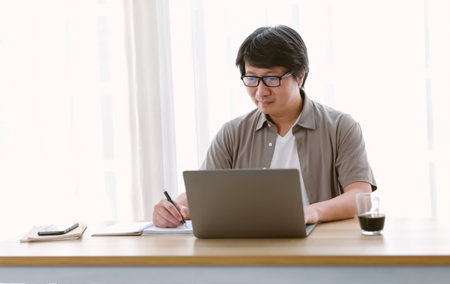 Mature Asian business man writing notes while working on laptop computer at his desk in home office....