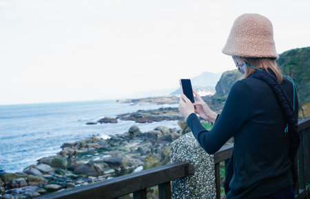 Back view portrait of Woman with face mask taking photo of a landscape mountains and blue lake. New normal lifestyle, travel and hobby conceptの写真素材