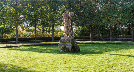 Old antique angel statue on a green lawn surrounded by trimmed linden treesの写真素材
