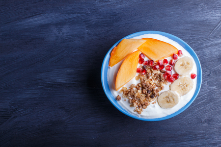 A blue plate with greek yogurt, granola, persimmon, banana, pomegranate black wooden background. top view. copy space.の写真素材