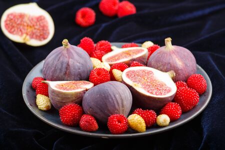 Fresh figs, strawberries and raspberries on blue ceramic plate on black concrete background and blue velvet textile. side view, close up, selective focusの写真素材