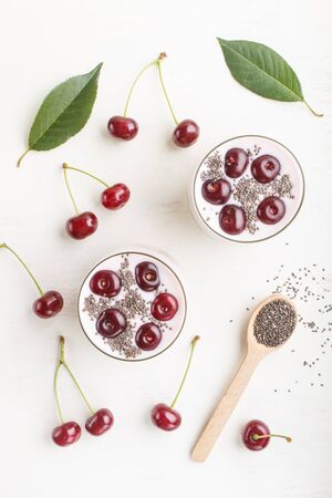 Yoghurt with cherries, chia seeds and granola in glass with wooden spoon on white wooden background. top view, flat lay.の写真素材