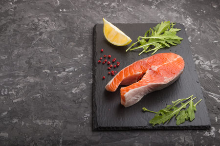 Raw salmon steak with arugula and lemon on a black slate board on a black concrete background. Side...
