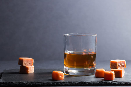 Glass of amber whiskey with caramel candies on a black stone slate board on blackの写真素材