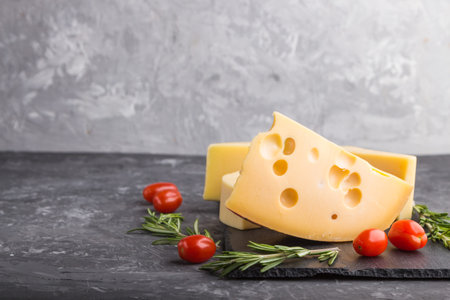 Various types of cheese with rosemary and tomatoes on black slate board on a black concrete...