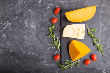 Various types of cheese with rosemary and tomatoes on black slate board on a black concrete...