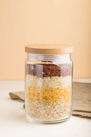glass jar with different kinds of rice poured in layers on white and orange background. side view, close up. Healthy food concept.の写真素材