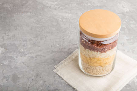 glass jar with different kinds of rice poured in layers on a gray concrete background. side view, close up, copy space. Healthy food concept.の写真素材
