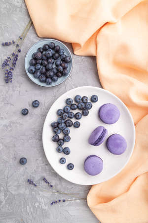 Purple macarons or macaroons cakes with blueberries on white ceramic plate on a gray concrete background and orange textile. Flat lay, top view, close up.の写真素材