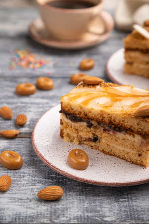 Honey cake with milk cream, caramel, almonds and a cup of coffee on a gray wooden background and linen textile. Side view, close up, selective focus.の写真素材