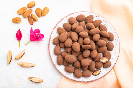 Almond in chocolate dragees on ceramic plate and a cup of coffee on white concrete background and...