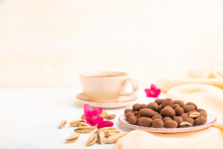 Almond in chocolate dragees on ceramic plate and a cup of coffee on white concrete background and...