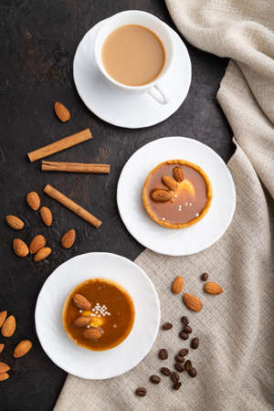 Sweet tartlets with almonds and caramel cream with cup of coffee on a black concrete background and linen textile. top view, flat lay, close up.の写真素材