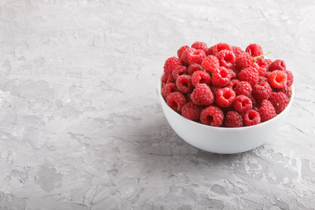 Fresh raspberry in white bowl on gray concrete background. side view, copy space.