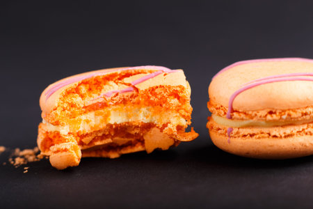 Whole and bitten orange macarons or macaroons cakes on black background. side view, close up, macro,...