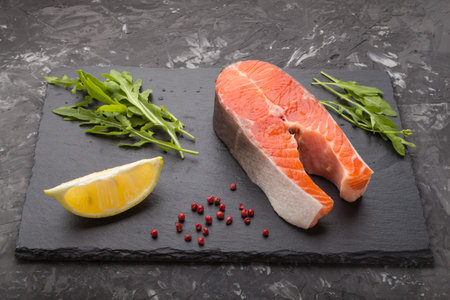 Raw salmon steak with arugula and lemon on a black slate board on a black concrete background. Side...