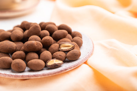 Almond in chocolate dragees on ceramic plate and a cup of coffee on white concrete background and...
