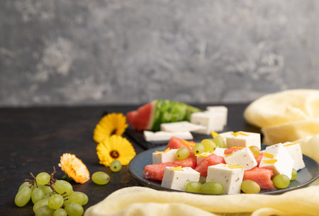 Vegetarian salad with watermelon, feta cheese, and grapes on blue ceramic plate on black concrete...