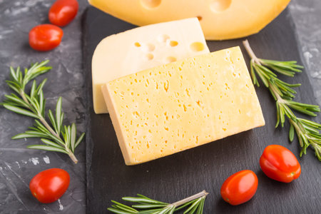 Various types of cheese with rosemary and tomatoes on black slate board on a black concrete background. Side view, close up, selective focus.の写真素材