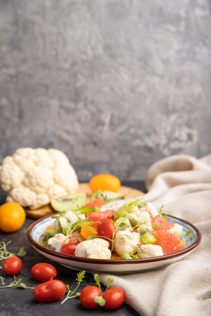Vegetarian salad of cauliflower cabbage, kiwi, tomatoes, microgreen sprouts on black concrete background and linen textile. Side view, copy space.の写真素材