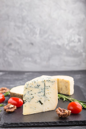 Blue cheese and various types of cheese with rosemary and tomatoes on black slate board on a black concrete background. Side view, copy space, selective focus.の写真素材