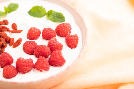 Yogurt with raspberry and goji berries in ceramic bowl on white concrete background and orange linen...