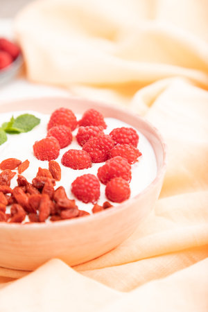 Yogurt with raspberry and goji berries in ceramic bowl on white concrete background and orange linen...