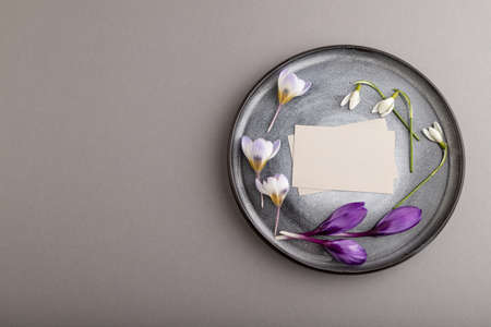 Gray paper invitation card, mockup with crocus and galanthus flowers on ceramic plate and gray background. Blank, top view, flat lay, still life, copy space, wedding invitation.の写真素材