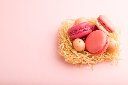 Purple and pink macaroons and chocolate eggs in decorative paper nest on pink pastel background....