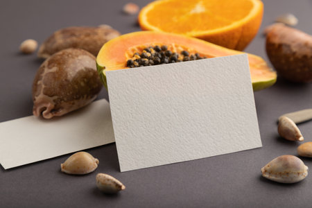 White paper business card with ripe cut papaya, orange, seashells on gray pastel pastel background. Side view, copy space. Tropical, healthy food, vacation, holidays concept.の写真素材