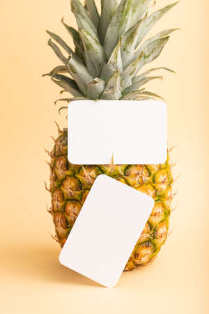 White paper business card with ripe pineapple on orange pastel background. Side view, copy space. Tropical, healthy food, vacation, holidays concept.の写真素材