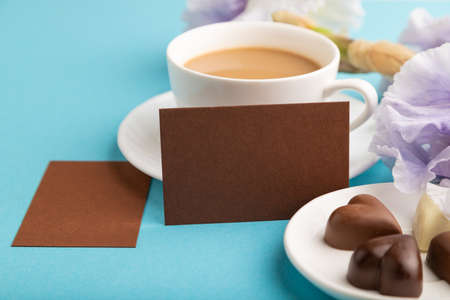 Brown business card with cup of cioffee, chocolate candies and iris flowers on blue pastel background. side view, copy space, still life. Breakfast, morning, spring concept.の写真素材