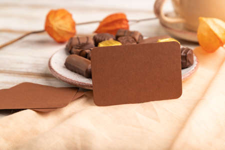 Brown business card with chocolate candies and physalis flowers on a white wooden background and orange textile. side view, close up, selective focus.の写真素材