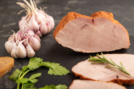Smoked pork ham on cutting slate board with pepper, garlic and herbs on black concrete background. Side view, close up, selective focus.の写真素材