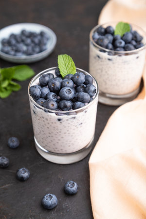 Yogurt with blueberry and chia in glass on black concrete background and orange linen textile. Side view, close up.