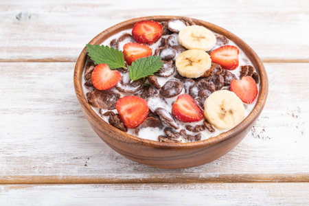 Chocolate cornflakes with milk and strawberry in wooden bowl on white wooden background. side view,...