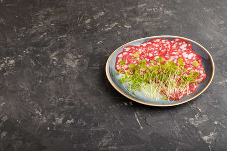 Slices of smoked cervelat salami sausage with spinach microgreen on black concrete background. Side view, copy space.の写真素材