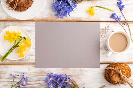 Gray paper sheet with oatmeal cookies, spring snowdrop flowers bluebells, narcissus and cup of coffee on white wooden background. top view, flat lay, copy space, still life. Breakfast, morning, spring concept.の写真素材