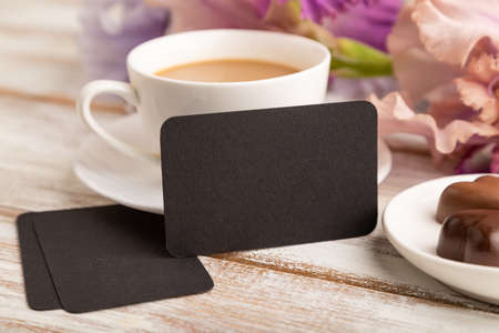 Black business card with cup of cioffee, chocolate candies and iris flowers on white wooden background. side view, copy space, still life. Breakfast, morning, spring concept.の写真素材