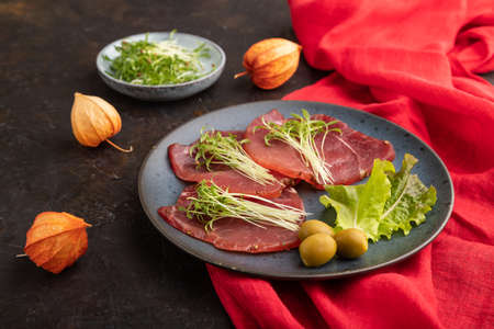 Slices of smoked salted meat with cilantro microgreen on black concrete background and red textile. side view, close up.の写真素材