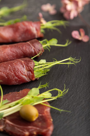 Slices of smoked salted meat with green pea microgreen on black concrete background. Side view,...